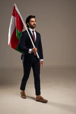 Celebrating the anniversary of Oman's National Day, the concept of love for the homeland and belonging to it, participation in national holidays and events, pride and honor in the symbols and flag of Oman, a portrait of a young Arab Gulf Omani man wearing a formal suit holding the flag of the Sultanate of Oman in his hand, full-body, white background.