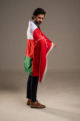 Pride in the symbols and the Omani flag, celebrating the anniversary of the Omani National Day, the concept of love for the homeland and belonging to it, participation in national holidays and events, a portrait of a young Arab Gulf Omani man wearing a formal suit with the flag of the Sultanate of Oman draped over his shoulder, full-body, white background.
