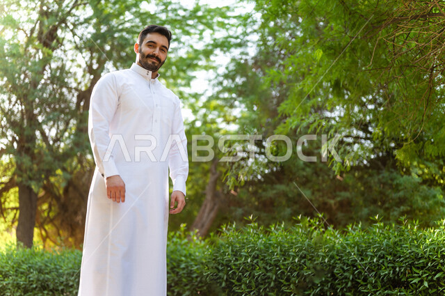 صورة لرجل عربي سعودي خليجي أنيق ، يرتدي الزي التقليدي السعودي ، رجل سعودي يتنزه في إحدى الحدائق الترفيهيه الرائعة بالمملكة العربية السعودية في وضع النهار ، خلفية للطبيعة في السعودية ، جمال المناظر الطبيعية الخلابة في السعودية