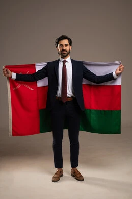Participation in national holidays and events, love for the homeland and belonging to it, pride in the symbols and the Omani flag, celebrating the anniversary of the Omani National Day, a portrait of a young Arab Gulf Omani man wearing a formal suit holding the flag of the Sultanate of Oman in his hand, full-body, white background.