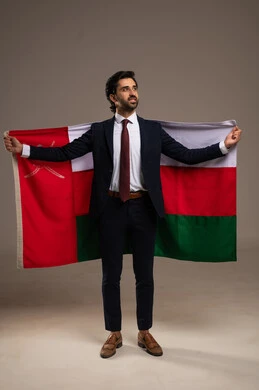 The concept of participation in national holidays and events, love for the homeland and belonging to it, pride in the symbols and the Omani flag, celebrating the anniversary of the Omani National Day, a portrait of a young Arab Gulf Omani man wearing a formal suit holding the flag of the Sultanate of Oman in his hand, full-body, white background.