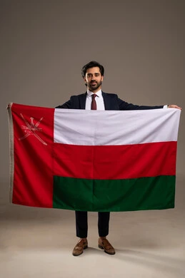 Participation in national holidays and events, love for the homeland and belonging to it, pride in the symbols and the Omani flag, celebrating the anniversary of the Omani National Day, a portrait of a young Arab Gulf Omani man wearing a formal suit holding the flag of the Sultanate of Oman in his hand, full-body, white background.