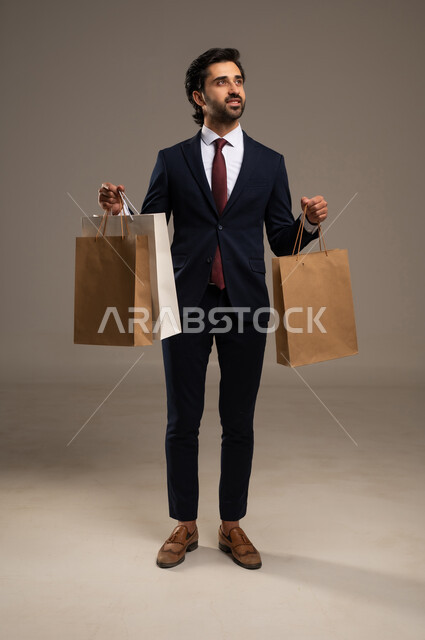 Shopping and taking the necessary supplies, benefiting from discount and clearance seasons, commercial purchases and gifts, a portrait of a Saudi Arabian Gulf man wearing official attire and holding paper bags in his hand, full body image, white background.