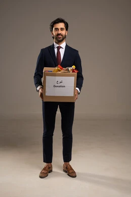 The concept of charity is spending in the way of God, caring about external appearance and self-confidence. A portrait of a Saudi Gulf Arab businessman wearing a formal suit, standing while holding a donation box and looking at the camera with a smile that expresses joy, with a white background.