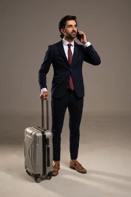 A smartphone and pulling a wheeled suitcase, looking at the phone to check flight details or time, paying attention to appearance and self-confidence, a portrait of a Saudi Gulf Arab businessman wearing a formal suit standing and holding a mobile phone with a suitcase beside him, white background.