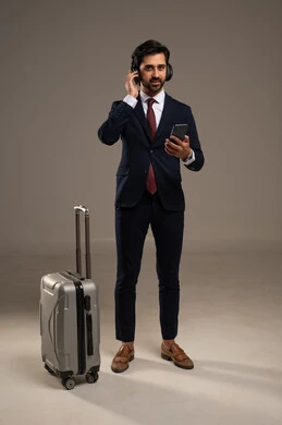 استخدام التكنولوجيا في التواصل قبل السفر، الاهتمام بالمظهر الخارجي والثقة بالنفس، بورتريه لرجل أعمال عربي خليجي سعودي يرتدي البدلة الرسمية يقف ويضع سماعات الرأس ويحمل هاتف محمول وبجانبه حقيبة سفر,  خلفية بيضاء