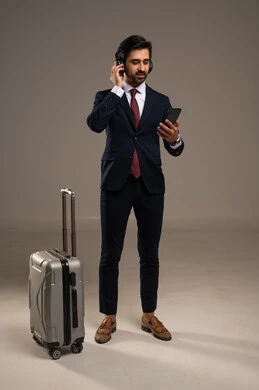 إجراء مكالمات فيديو أو اجتماعات عبر الإنترنت قبل السفر، الاهتمام بالمظهر الخارجي والثقة بالنفس، بورتريه لرجل أعمال عربي خليجي سعودي يرتدي البدلة الرسمية يقف ويضع سماعات الرأس ويحمل هاتف محمول وبجانبه حقيبة سفر,  خلفية بيضاء