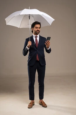 أجواء شتوية، مفهوم الحماية من المطر, استخدام التقنيات الحديثة، بورتريه لرجل أعمال عربي خليجي سعودي يرتدي البدلة الرسمية يقف ويحمل مظلة وهاتف محمول, الاهتمام بالمظهر الخارجي، خلفية بيضاء