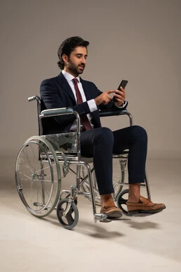 Conducting a video phone call, managing and organizing business affairs through technical devices, integrating work with technology and tech, a portrait of an Arab Gulf Saudi man with special needs wearing formal attire sitting in a wheelchair holding a mobile phone, white background.