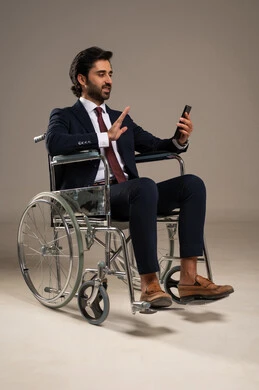 Conducting a video phone call, managing and organizing business affairs through technical devices, integrating work with technology and tech, a portrait of an Arab Gulf Saudi man with special needs wearing formal attire sitting in a wheelchair holding a mobile phone, white background.