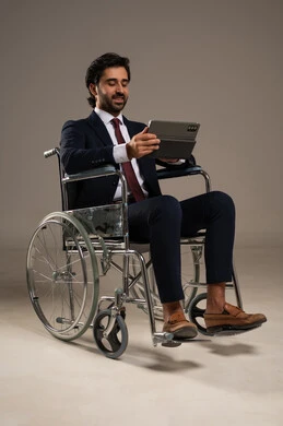 Managing and organizing business affairs through technical devices, integrating work with technology and tech, a portrait of a Saudi Arabian Gulf man with special needs wearing formal attire sitting in a wheelchair working on a tablet, white background.