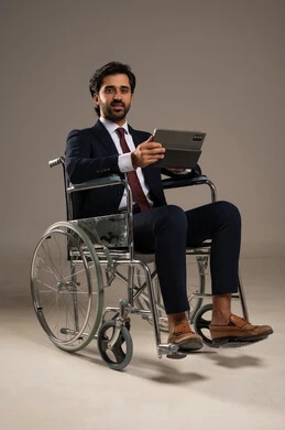 إدارة شؤون الأعمال وتنظيمها عبر الاجهزة التقنية، دمج العمل مع التقنية والتكنولوجيا، بورتريه لرجل عربي خليجي سعودي من ذوي الاحتياجات الخاصة يرتدي الزي الرسمي يجلس على كرسي متحرك يعمل على جهاز التابليت , خلفية بيضاء