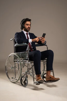 استخدام التكنولوجيا و التقنيات الحديثة، أسلوب حياة ذوي الهمم، الدردشة والتواصل مع الأهل والأصدقاء، بورتريه لرجل عربي خليجي سعودي مقعد يرتدي البدلة الرسمية يجلس على كرسي متحرك يحمل بيده الهاتف المحمول، صورة لكامل الجسم، خلفية بيضاء
