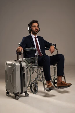 البحث والاستكشاف حول العالم, الاستعداد لقضاء وقت ممتع في الرحلات السياحية, بورتريه لرجل عربي خليجي سعودي مبتسم يرتدي البدلة الرسمية يجلس على الكرسي المتحرك يضع سماعات الرأس وبجانبه حقيبة الامتعة , الاستمتاع بسماع الاغاني المفضلة في وقت السفر، خلفية بيضاء