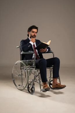 قضاء وقت الفراغ بالتعلم والثقافة، مفهوم القراءة والمطالعة والتثقف,  حب المطالعة و الاستكشاف , ذوي الإحتياجات الخاصة والهمم, بورتريه لشاب عربي خليجي سعودي يرتدي البدلة الرسمية يجلس على كرسي متحرك ويمسك بيديه كتاب, خلفية بيضاء
