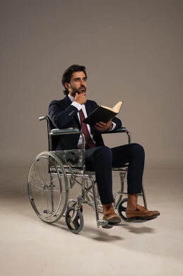 The love of reading and exploration, spending free time learning and gaining knowledge, the concept of reading and literacy, people with special needs and determination, a portrait of a young Arab Gulf Saudi man wearing a formal suit sitting in a wheelchair holding a book in his hands, white background.
