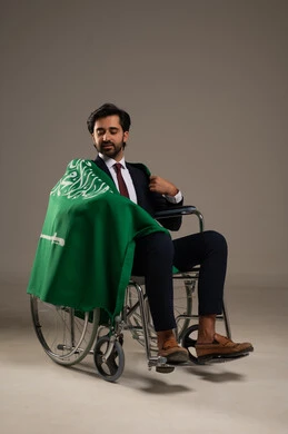 Pride in Saudi identity, participation in national events on September 23 and commemorating National Day, a portrait of a smiling Arab Saudi Gulf man wearing a formal suit sitting in a wheelchair and draping the flag of the Kingdom over his shoulders, the anniversary of Saudi Flag Day on March 11, white background.