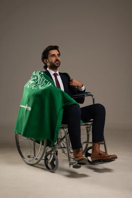 المشاركة في المناسبات الوطنية في 23 سبتمبر واستذكار اليوم الوطني, الفخر بالهوية السعودية, بورتريه  لرجل عربي سعودي خليجي مبتسم يرتدي البدلة الرسميةيجلس على كرسي متحرك ويضع على اكتافه علم المملكة, ذكرى يوم العلم السعودي 11 مارس, خلفية بيضاء