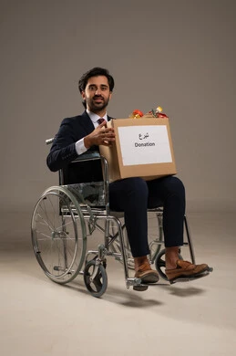 Humanitarian aid and donations, encouraging charitable Islamic work, feeling for others and helping them, a portrait of a young Arab Gulf Saudi man with special needs wearing a formal suit sitting in a wheelchair holding a donation box, white background.
