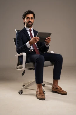 التسوق عبر الانترنت باستخدام التابلت, قضاء الوقت بتصفح برامج التواصل الاجتماعي, إنجاز المهام عن بعد،  استعمال أجهزة تقنية حديثة، بورتريه مقرب لرجل عربي سعودي خليجي يرتدي البدلة الرسمية يجلس على كرسي مريح ويستخدم الجهاز اللوحي، خلفية بيضاء