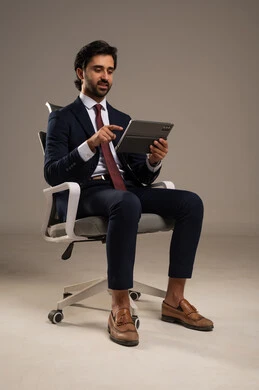 قضاء الوقت بتصفح برامج التواصل الاجتماعي, إنجاز المهام عن بعد، التسوق عبر الانترنت باستخدام التابلت, استعمال أجهزة تقنية حديثة، بورتريه مقرب لرجل عربي سعودي خليجي يرتدي البدلة الرسمية يجلس على كرسي مريح ويستخدم الجهاز اللوحي، خلفية بيضاء