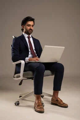 مفهوم العمل عن بعد,  انجاز وادارة المهام عبر الأونلاين, دمج العمل مع التقنية والتكنولوجيا الحديثة والمتطورة, بورتريه لشاب عربي خليجي سعودي يرتدي البدلة الرسمية و يستخدم الحاسب المحمول, يجلس على كرسي مريح, استخدام الأجهزة والتقنيات الحديثة، خلفية بيضاء