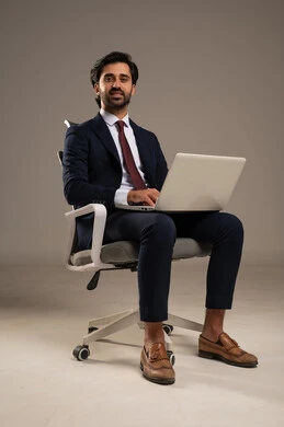 دمج العمل مع التقنية والتكنولوجيا الحديثة والمتطورة, انجاز وادارة المهام عبر الأونلاين, مفهوم العمل عن بعد, بورتريه لشاب عربي خليجي سعودي يرتدي البدلة الرسمية و يستخدم الحاسب المحمول, يجلس على كرسي مريح, استخدام الأجهزة والتقنيات الحديثة، خلفية بيضاء
