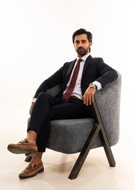 الجدية والحدة في النظر، إيماءات القوة والثقة، مودل لعرض الملابس والاكسسوارات، مفهوم الاناقة والجمال، مفهوم الشكر والامتنان، بورتريه لشاب عربي خليجي سعودي يرتدي الطقم الرسمي يجلس على اريكة مريحة باللون الرمادي بثقة وعزة نفس، خلفية بيضاء
