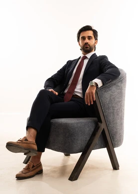 A look and gestures of power and confidence, a model for showcasing clothing and accessories, the concept of elegance and beauty, the concept of gratitude and appreciation, attention to health and physical fitness, a portrait of a young Arab Gulf Saudi man wearing formal attire sitting on a comfortable gray sofa with confidence and self-esteem, white background.