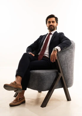 Masculinity, elegance, and confidence, a model for showcasing clothing and accessories, the concept of elegance and beauty, the concept of gratitude and appreciation, attention to health and physical fitness, a portrait of a young Arab Gulf Saudi man wearing a formal suit sitting on a comfortable gray sofa with confidence and dignity, white background.