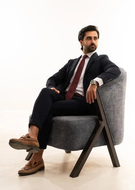 A model for showcasing clothing and accessories, gestures of strength and confidence, the concept of elegance and beauty, the concept of gratitude and appreciation, attention to health and physical fitness, a portrait of a young Arab Gulf Saudi man wearing formal attire sitting on a comfortable gray sofa with confidence and self-esteem, white background.