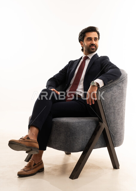 A model man showcasing clothing and accessories, gestures of strength and confidence, the concept of elegance and beauty, the concept of gratitude and appreciation, attention to health and physical fitness, a portrait of a young Arab Gulf Saudi man wearing a formal suit sitting on a comfortable gray sofa with confidence and self-esteem, white background.