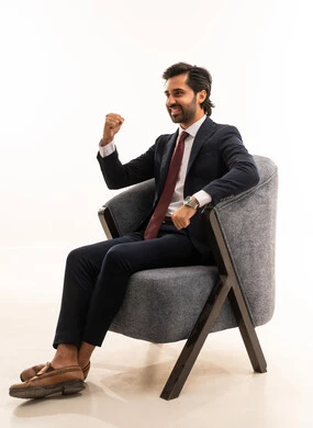 رفع قبضة اليد لتسجل هدف، التفاعل والاندماج مع المباريات، رجل سعودي يقوم بتشجيع المنتخب الوطني,  بورتريه لشاب عربي خليجي سعودي يرتدي الطقم الرسمي ويجلس على اريكة مريحة بايماءات السرور من تسجيل الاهداف,  مفهوم الجمال والاناقة والرجولة، خلفية بيضاء