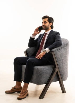 مكالمة هاتفية باستخدام الموبايل، الدردشة والتواصل مع الاصدقاء بإيماءات السعادة والفرح، بورتريه لشاب عربي خليجي سعودي يرتدي البدلة الرسمية يجلس على اريكة مريحة باللون الرمادي يمسك بيده الهاتف المحمول ،رجل سعودي يستخدم الأجهزة والتقنيات الحديثة،  خلفية بيضاء