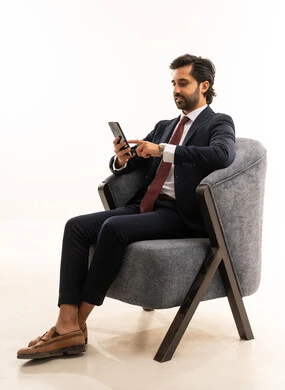 الدردشة والتواصل مع الاصدقاء بإيماءات السعادة والفرح، بورتريه لشاب عربي خليجي سعودي يرتدي البدلة الرسمية يجلس على اريكة مريحة باللون الرمادي يمسك بيده الهاتف المحمول ،رجل سعودي يستخدم الأجهزة والتقنيات الحديثة،  خلفية بيضاء