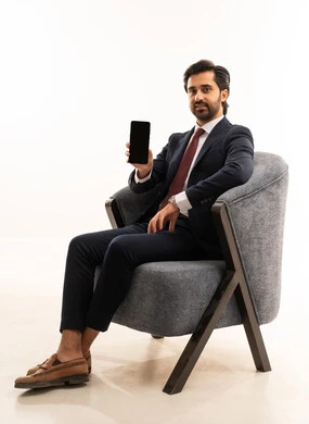 A black screen on the mobile phone, chatting and communicating with friends through gestures of happiness and joy, a portrait of a young Arab Gulf Saudi man wearing a formal suit sitting on a comfortable gray sofa holding a mobile phone in his hand, a Saudi man using modern devices and technologies, white background.