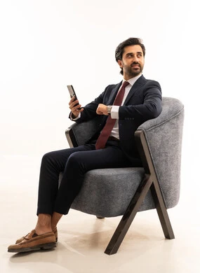 رجل سعودي يستخدم الأجهزة والتقنيات الحديثة، تصفح مواقع التواصل الاجتماعي، الدردشة والتواصل مع الاصدقاء بإيماءات السعادة والفرح، بورتريه لشاب عربي خليجي سعودي يرتدي البدلة الرسمية يجلس على اريكة مريحة باللون الرمادي يمسك بيده الهاتف المحمول ، خلفية بيضاء