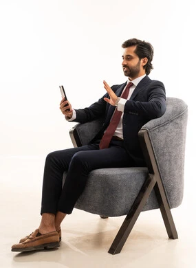 اجراء مكالمة فيديو على الموبايل، الدردشة والتواصل مع الاصدقاء بإيماءات السعادة والفرح، بورتريه لشاب عربي خليجي سعودي يرتدي البدلة الرسمية يجلس على اريكة مريحة باللون الرمادي يمسك بيده الهاتف المحمول ،رجل سعودي يستخدم الأجهزة والتقنيات الحديثة،  خلفية بيضاء