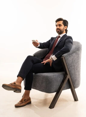 Following the latest news on the television screen, using a remote control to flip between channels, a Saudi man using modern devices and technologies, browsing social media, a portrait of a young Arab Gulf Saudi man wearing a formal suit sitting on a comfortable gray sofa, with a white background.