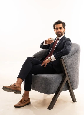 استخدام جهاز تحكم للتقليب بين القنوات، متابعة الاخبار الجديدة على شاشة التلفاز، رجل سعودي يستخدم الأجهزة والتقنيات الحديثة، تصفح مواقع التواصل الاجتماعي، بورتريه لشاب عربي خليجي سعودي يرتدي البدلة الرسمية يجلس على اريكة مريحة باللون الرمادي ، خلفية بيضاء