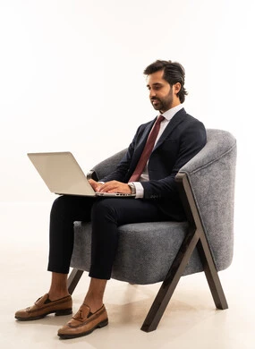 مهارة الكتابة السريعة على لوحة التحكم، انهاء مهام العمل عن بعد، رجل سعودي يستخدم الأجهزة والتقنيات الحديثة، تصفح مواقع التواصل الاجتماعي، بورتريه لشاب عربي خليجي سعودي يرتدي البدلة الرسمية يجلس على اريكة مريحة باللون الرمادي يمسك بيده الحاسب المحمول ، خلفية بيضاء