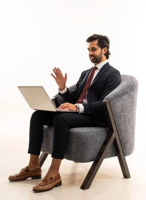 القاء التحيو عبر كالمة فيديو، انهاء مهام العمل عن بعد، رجل سعودي يستخدم الأجهزة والتقنيات الحديثة، تصفح مواقع التواصل الاجتماعي، بورتريه لشاب عربي خليجي سعودي يرتدي البدلة الرسمية يجلس على اريكة مريحة باللون الرمادي يمسك بيده الحاسب المحمول ، خلفية بيضاء