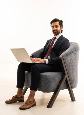 انهاء مهام العمل عن بعد، رجل سعودي يستخدم الأجهزة والتقنيات الحديثة، تصفح مواقع التواصل الاجتماعي، بورتريه لشاب عربي خليجي سعودي يرتدي البدلة الرسمية يجلس على اريكة مريحة باللون الرمادي يمسك بيده الحاسب المحمول ، خلفية بيضاء