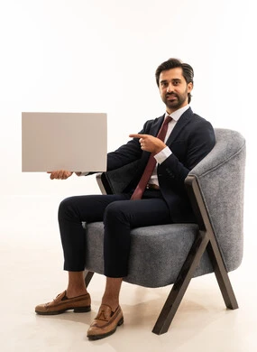 Using the index and pointer fingers for the board, advertising and marketing campaigns, white rectangular mockup, the concept of elegance, beauty, and masculinity, recreational youth activities, a portrait of a young Arab Gulf Saudi man wearing formal attire sitting on a comfortable sofa holding a white rectangular board, white background.