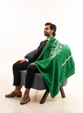 الاحتفال باليوم الوطني السعودي، أعيادومناسبات وطنية، رجل سعودي يقوم بالاحتفال باليوم الوطني,  بورتريه لشاب عربي خليجي سعودي يرتدي الطقم الرسمي ويجلس على اريكة مريحة ويضع على كتفه علم المملكة بايماءات السرور,  مفهوم الجمال والاناقة والرجولة، خلفية بيضاء