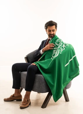 رجل سعودي يقوم بالاحتفال باليوم الوطني, أعياد ومناسبات وطنية، الاحتفال باليوم الوطني السعودي،  بورتريه لشاب عربي خليجي سعودي يرتدي الطقم الرسمي ويجلس على اريكة مريحة ويضع على كتفه علم المملكة بايماءات السرور,  مفهوم الجمال والاناقة والرجولة، خلفية بيضاء