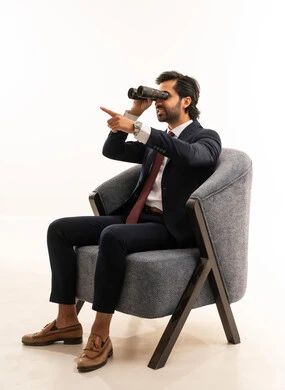 Pointing to something, the concept of exploration and adventure, searching for something and expanding the field of vision using binoculars, a portrait of a young Arab Gulf Saudi man wearing formal attire sitting on a comfortable sofa and looking through binoculars, a Saudi man searching and exploring, white background.