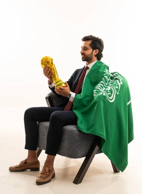تامل تفاصيل الكأس الذهبي، رجل سعودي يقوم بتشجيع المنتخب الوطني, التفاعل والاندماج مع المباريات،  بورتريه لشاب عربي خليجي سعودي يرتدي الطقم الرسمي ويجلس على اريكة مريحة علم المملكة على كتفه ويحمل كأس ذهبي, خلفية بيضاء