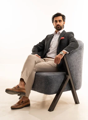 ساعة يد باهظة الثمن، إيماءات القوة والثقة، مفهوم الاناقة والجمال، مفهوم الشكر والامتنان، الاهتمام بالصحة واللياقة الجسدية، بورتريه لشاب عربي خليجي سعودي يرتدي الطقم الرسمي يجلس على اريكة مريحة باللون الرمادي بثقة وعزة نفس، خلفية بيضاء