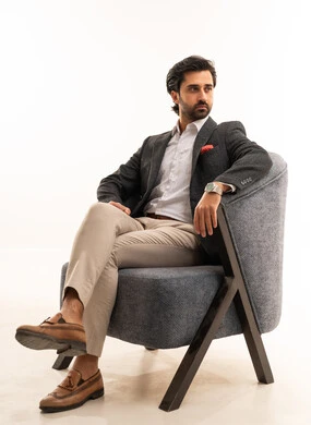 حذاء جلد بني ، إيماءات القوة والثقة، مفهوم الاناقة والجمال، مفهوم الشكر والامتنان، الاهتمام بالصحة واللياقة الجسدية، بورتريه لشاب عربي خليجي سعودي يرتدي الطقم الرسمي يجلس على اريكة مريحة باللون الرمادي بثقة وعزة نفس، خلفية بيضاء